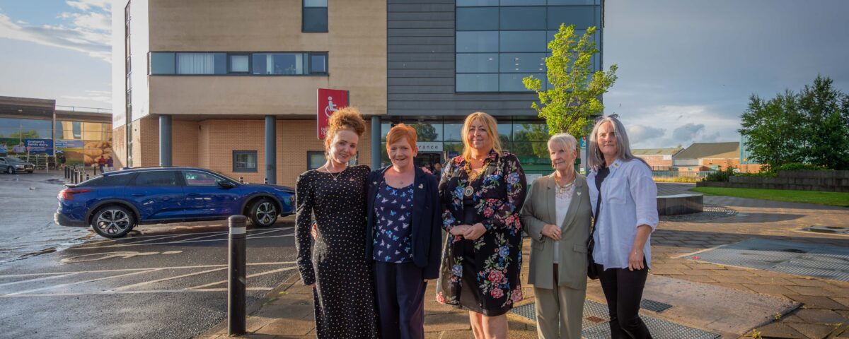 Five people stand in front of Tweedie Hall and Linwood Library. The group poses on a wet pavement, wearing casual and semi-formal attire. A blue car and a modern building can be seen in the background under a partly cloudy sky.
