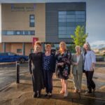 Five people stand in front of Tweedie Hall and Linwood Library. The group poses on a wet pavement, wearing casual and semi-formal attire. A blue car and a modern building can be seen in the background under a partly cloudy sky.