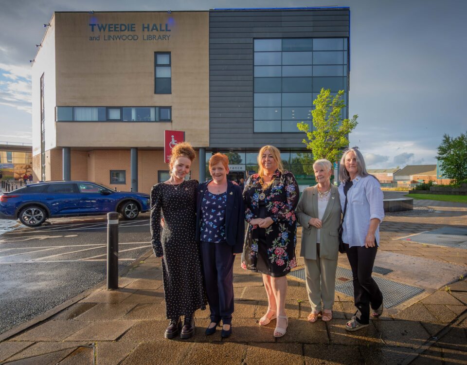 Five people stand in front of Tweedie Hall and Linwood Library. The group poses on a wet pavement, wearing casual and semi-formal attire. A blue car and a modern building can be seen in the background under a partly cloudy sky.