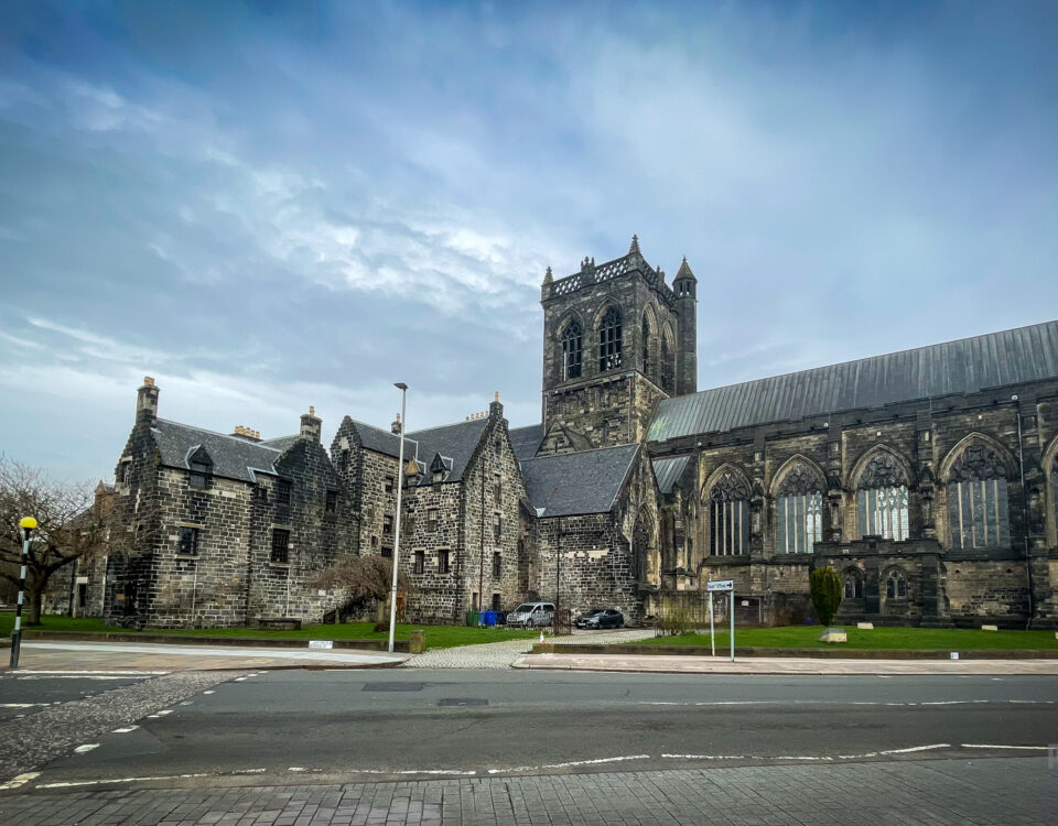 A large, historic stone cathedral with Gothic architecture stands under a cloudy sky. The building features intricate stained glass windows and a tall bell tower. A road with pedestrian crossings is in the foreground.