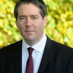 A man in a black suit and red tie is standing outdoors. The background features blurred greenery with yellow and green leaves. He has short dark hair and is looking directly at the camera.
