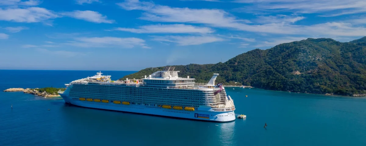 A large cruise ship is anchored in a calm blue sea with a mountainous island in the background under a partly cloudy sky. The ship is white with yellow lifeboats, and the surrounding water is a deep blue.