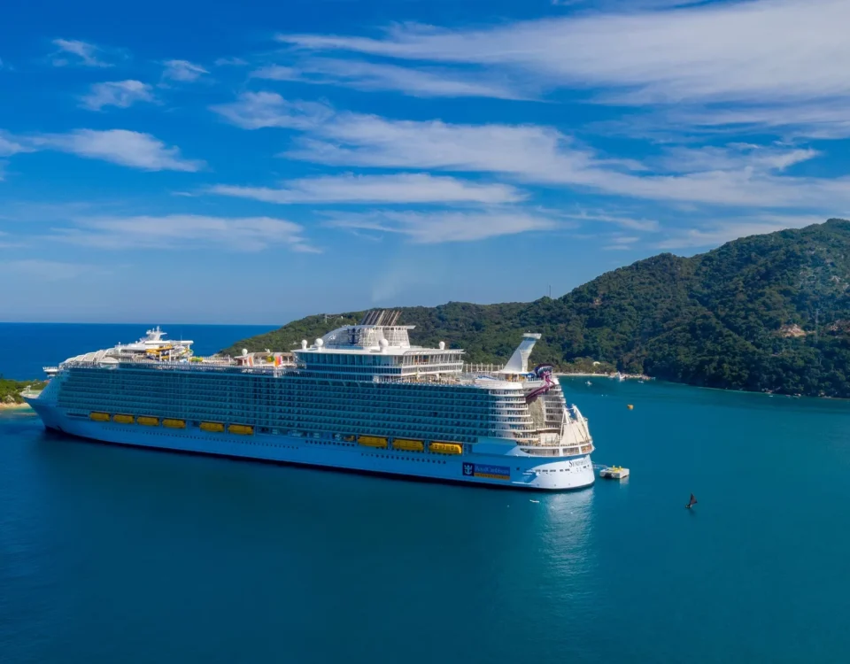 A large cruise ship is anchored in a calm blue sea with a mountainous island in the background under a partly cloudy sky. The ship is white with yellow lifeboats, and the surrounding water is a deep blue.