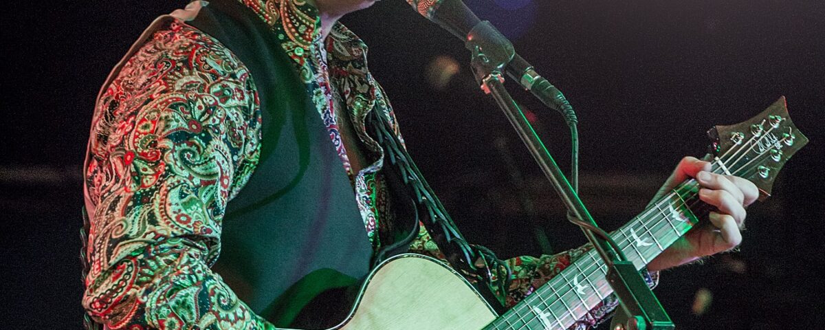 A musician passionately sings and plays an acoustic guitar on stage. He is wearing a colorful patterned shirt, vest, and cap, performing under bright stage lights.