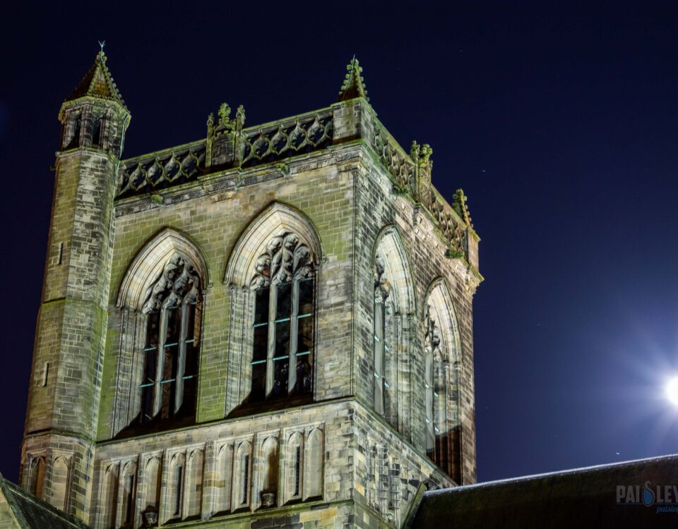 A night view of a Gothic-style cathedral tower with pointed arches and decorative stonework, illuminated against a dark sky. A bright full moon shines to the right of the tower.