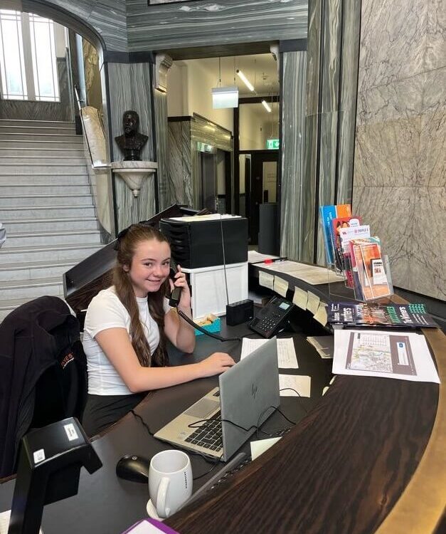 A person with long hair is sitting at a reception desk, talking on the phone. There is a laptop, phone, and brochures on the desk. The background shows a staircase and marble walls.
