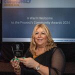 A person holding an award stands in front of a screen displaying "Provost’s Community Awards 2024." They're smiling, wearing a black outfit, and the event appears to be a formal awards ceremony.