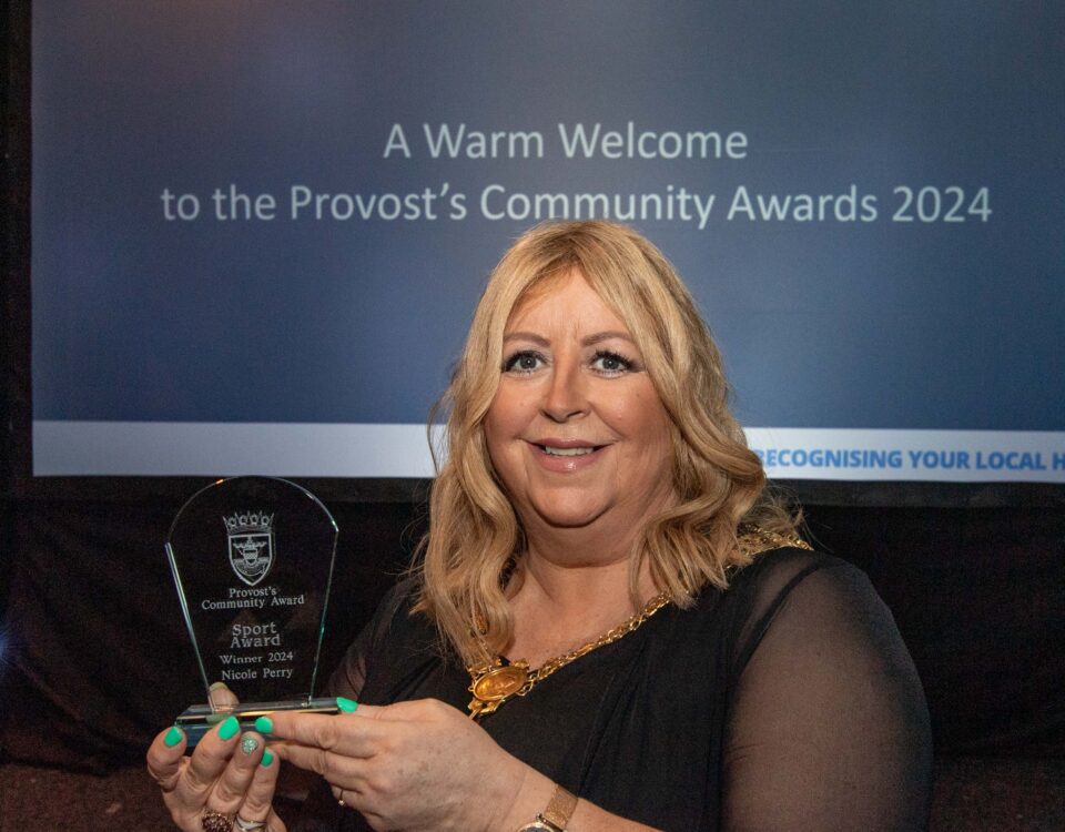 A person holding an award stands in front of a screen displaying "Provost’s Community Awards 2024." They're smiling, wearing a black outfit, and the event appears to be a formal awards ceremony.
