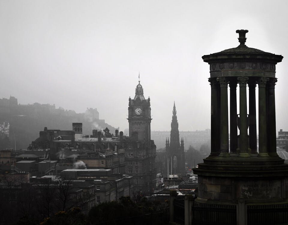 A moody, overcast view of a cityscape featuring a tall, cylindrical monument in the foreground. In the background, historic buildings and a clock tower rise through the mist, creating a dramatic silhouette against the gray sky.
