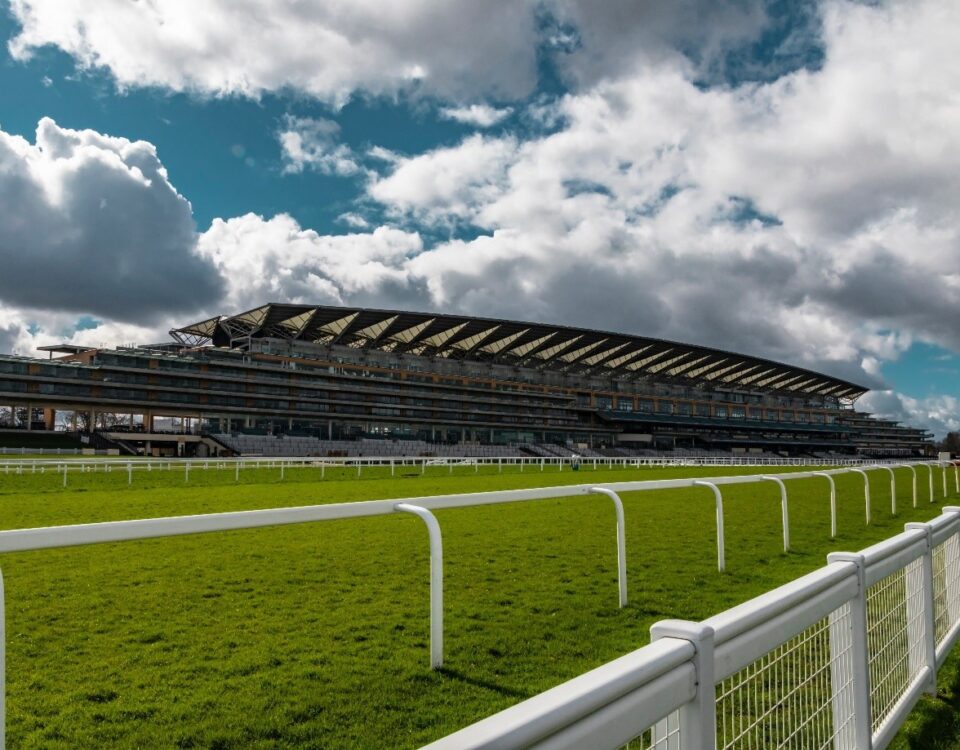 A grandstand at a racecourse under a partly cloudy sky, with a well-maintained grass track in the foreground. The structure extends horizontally, showcasing its modern architectural design.