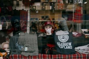 Store window display featuring a mannequin head with a hat and mask, surrounded by Glasgow-themed clothing and accessories. Items include sweaters, t-shirts, gloves, and patterned bags, all set against a tartan backdrop.