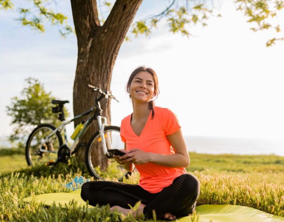 A woman in a bright orange shirt sits cross-legged on a yoga mat in a grassy area near a tree, holding a phone and smiling. A bicycle rests against the tree, with a backdrop of greenery and a sunlit sky.