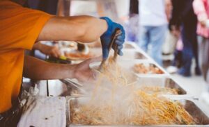 A person in an orange shirt and blue gloves serves steaming noodles from a large tray into a takeout container at a food stall. Other trays with various dishes are visible in the background, with people standing nearby.