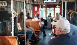 A group of people sit inside a trolley bus with wooden seats. A man in the foreground wears a white cap. The passengers are casually dressed, and a screen is visible at the front of the trolley. Sunlight filters in from the windows.