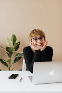 A smiling person with short hair and glasses sits at a desk with a laptop, phone, pen, and a potted plant in the background, resting their face in their hands and looking at the camera.