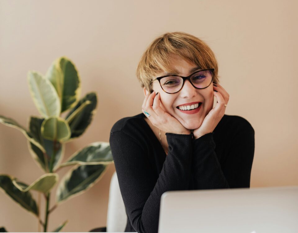 A smiling person with short hair and glasses sits at a desk with a laptop, phone, pen, and a potted plant in the background, resting their face in their hands and looking at the camera.