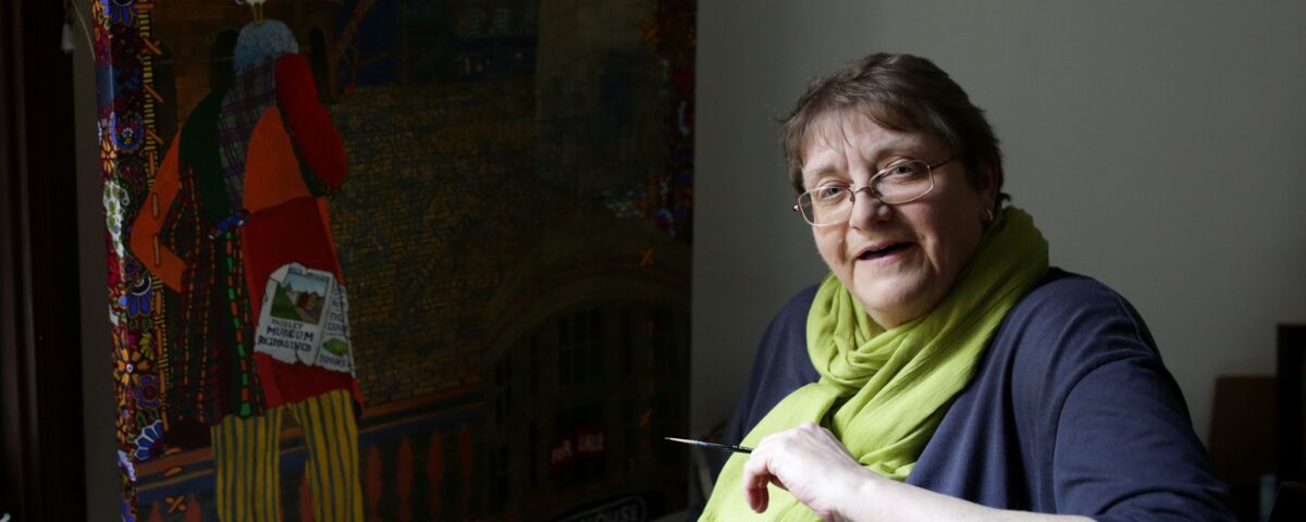 A woman with glasses and a green scarf smiles while sitting beside a colorful, detailed painting on an easel. She holds a paintbrush and is positioned near a window with natural light.