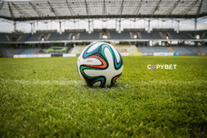 A soccer ball rests on the grass in the center of an empty football stadium, with rows of seats and a covered roof visible in the background. The word “COPYBET” appears to the right of the ball.