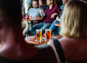 Two glasses of beer sit on a round wooden table in a busy, dimly lit bar. People are gathered in the background, chatting and laughing, creating a lively social atmosphere.