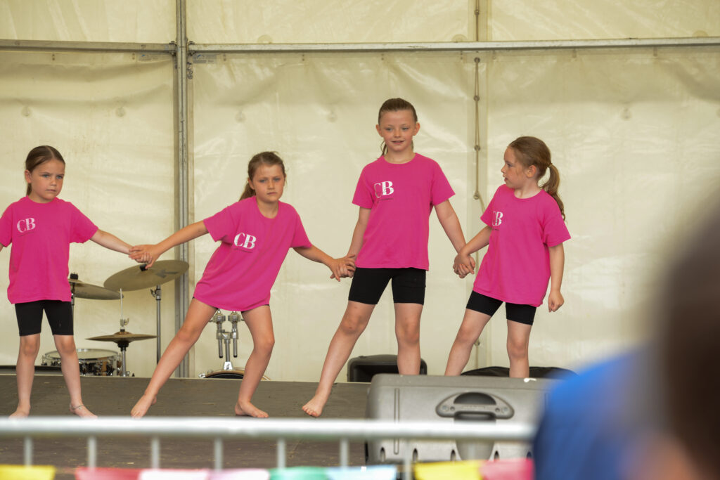 Four young children wearing matching pink shirts and black shorts are holding hands and performing a dance on a stage inside a tent. Drums and stage equipment are visible in the background.