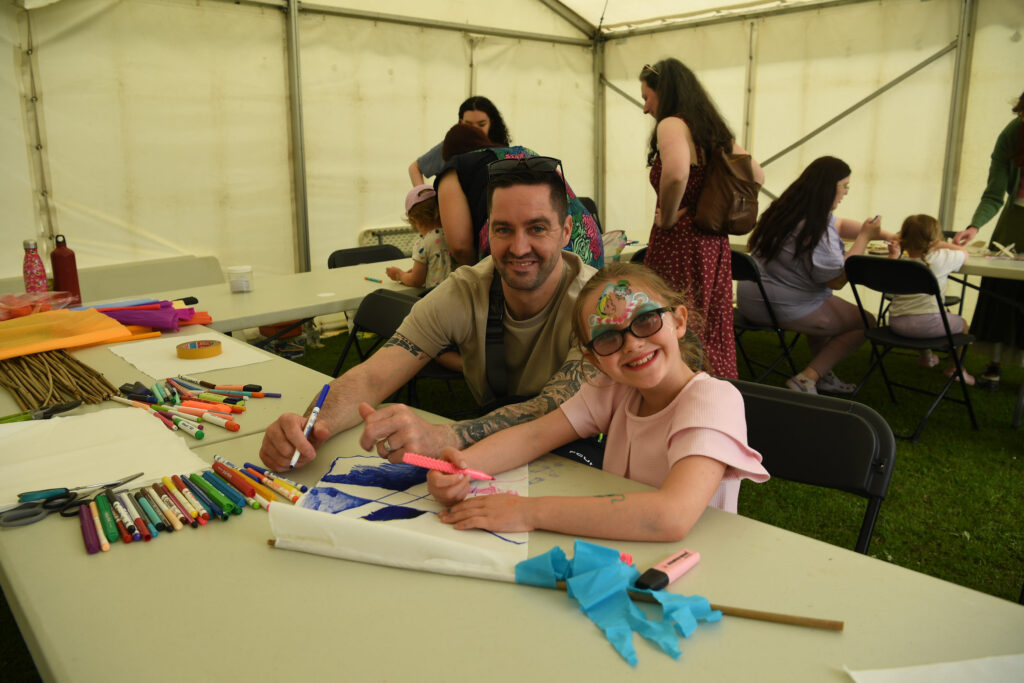 A smiling man and young girl with face paint sit at a table in a tent, drawing with markers and surrounded by art supplies. Other people are at tables in the background.