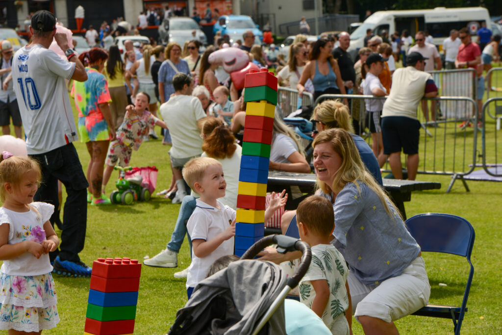 A woman and two young children laugh while playing with large colorful toy blocks at an outdoor event, surrounded by people enjoying activities in a park.