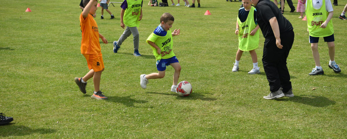 Children play soccer on a grassy field, wearing colorful jerseys. Spectators and parked cars are visible in the background. The scene is lively and takes place outdoors on a sunny day.