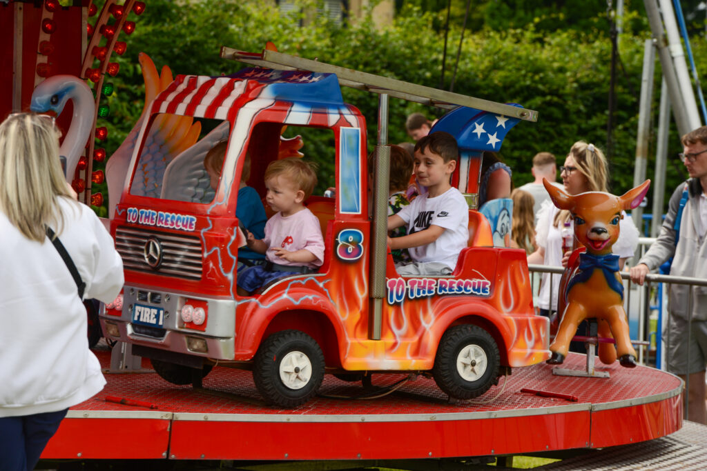 Children ride a colorful fire truck-themed carousel at an amusement park, accompanied by adults nearby. The ride features red flames, American flag motifs, and a reindeer figure at the front.