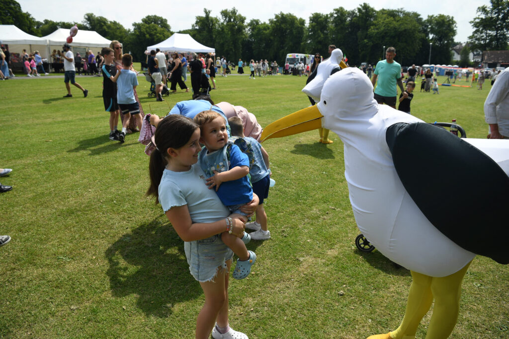 A young girl holds a toddler while standing on grass, facing a person dressed in a large bird costume. Other children and people are gathered outdoors at an event with tents and trees in the background.