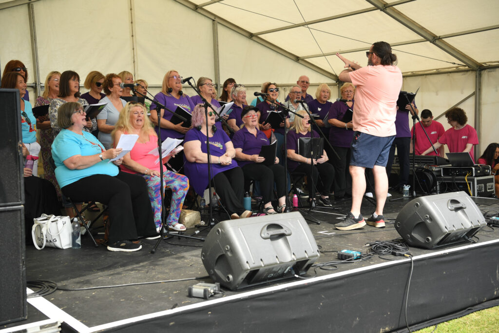 A choir of adults, mostly women, sit and stand in rows under a white tent, singing while a conductor in a pink shirt and shorts leads them; microphones and sound equipment are set up on stage.
