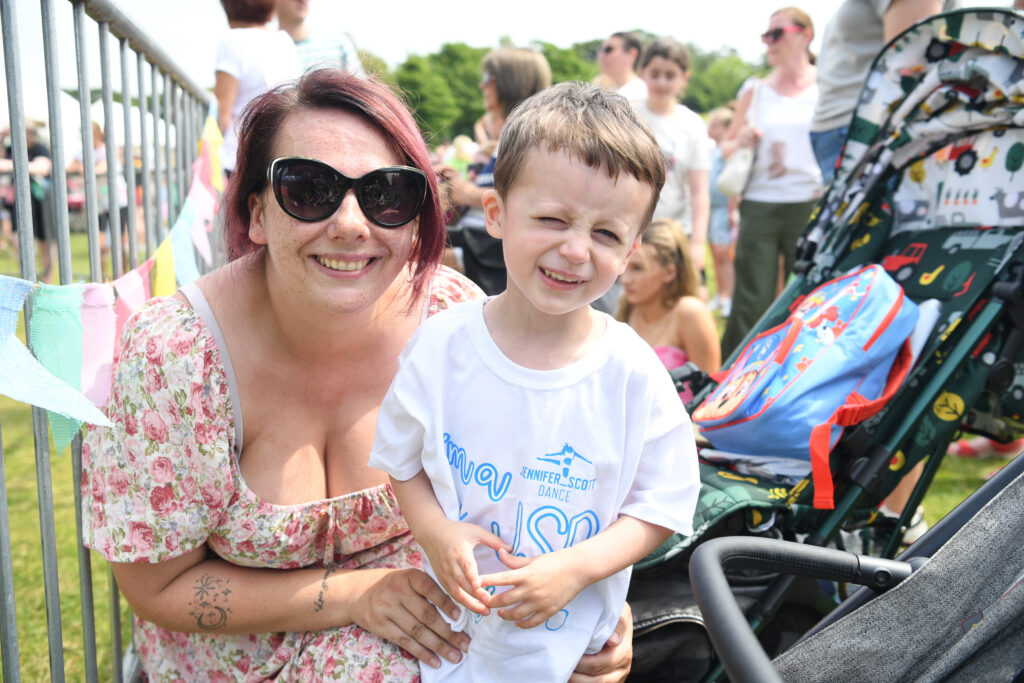 A woman with sunglasses kneels next to a smiling young boy in a white shirt. They are outdoors at a crowded event, with a stroller and colorful bunting nearby.