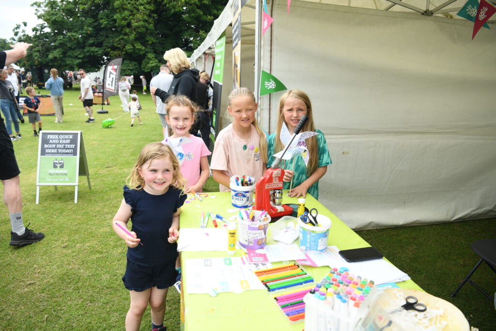Four young girls stand and smile at a craft table outdoors, covered with colorful markers and art supplies, in front of a white tent at a community event or festival. Other people and tents are visible in the background.