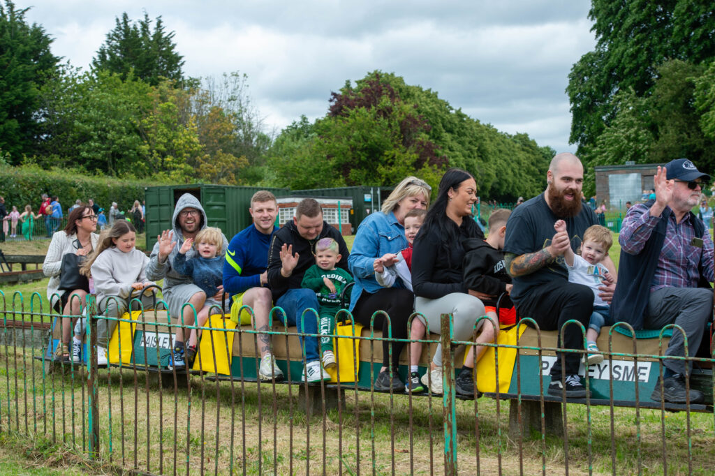 A group of adults and children sit on a small train ride outdoors, smiling and waving. The scene is in a park with green trees and partly cloudy skies in the background.
