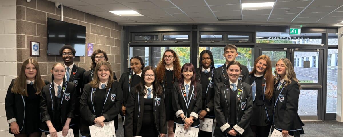 A group of students in school uniforms stand together indoors, smiling and holding papers. They are in a hallway with large windows and a brick wall, suggesting a school environment.