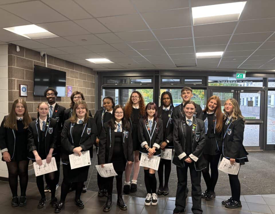A group of students in school uniforms stand together indoors, smiling and holding papers. They are in a hallway with large windows and a brick wall, suggesting a school environment.