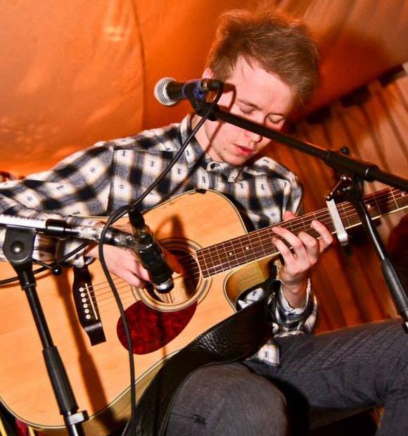 A young man in a plaid shirt sits and plays an acoustic guitar, focused on his performance, surrounded by microphones in a warmly lit setting.