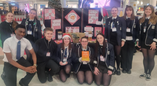 A group of students in school uniforms pose indoors in front of a festive display board. Some are kneeling, others stand behind. One student wears a Santa hat, and two hold an orange award plaque.