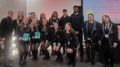 A group of students in school uniforms pose together indoors, some standing and others kneeling, in front of a presentation screen displaying colorful graphics. Two students hold up teal-colored booklets.