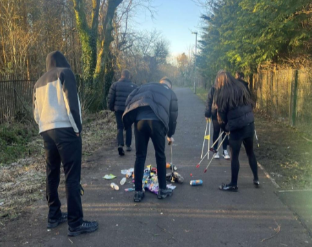 Five people in dark clothing are picking up litter with grabbers on a paved path lined with trees and a fence. Some trash is on the ground as they clean up the area on a sunny day.