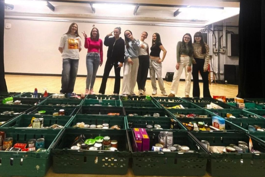 Eight people stand in a row behind green crates filled with various canned and packaged food items, in a large indoor room with wooden floors.