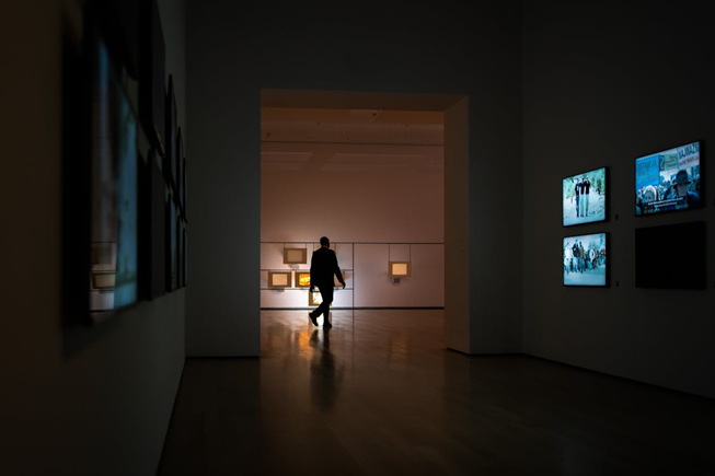 A person walks alone through a dimly lit art gallery, with illuminated displays on the walls and several screens showing images in the foreground. Shadows and warm light create a contemplative atmosphere.