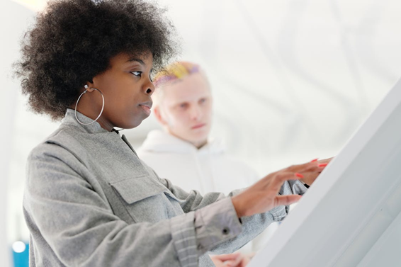 Two people use a large touchscreen monitor. The person in front, wearing a gray jacket and hoop earrings, interacts with the screen, while another person in a white hoodie stands behind them, watching.