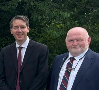 Two men in suits stand outdoors in front of green foliage, both smiling at the camera. The man on the left has short dark hair, while the man on the right has a bald head with a white beard and glasses hanging from his neck.