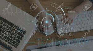 Hands using a computer mouse and keyboard next to a laptop on a wooden desk, with digital blue technology interface graphics and data charts overlaid on the image. The word "TECHNOLOGY" is displayed prominently.