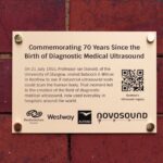 Plaque on a red brick wall commemorating 70 years since the birth of diagnostic medical ultrasound, detailing an event in 1955 and featuring logos, a QR code, and sponsor names.