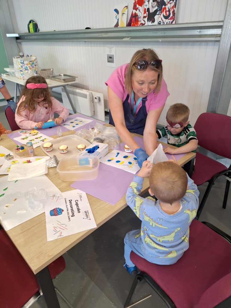 A woman helps three young children decorate cupcakes at a table covered with art supplies, paper, and a sign that reads "Cupcake Decorating." The kids are focused on their crafts and wearing colorful clothes.