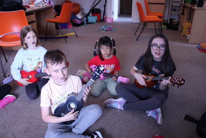 Four children sit on a carpeted floor in a classroom, each holding a ukulele. They appear to be learning or playing music together, with colorful chairs and various items in the background.