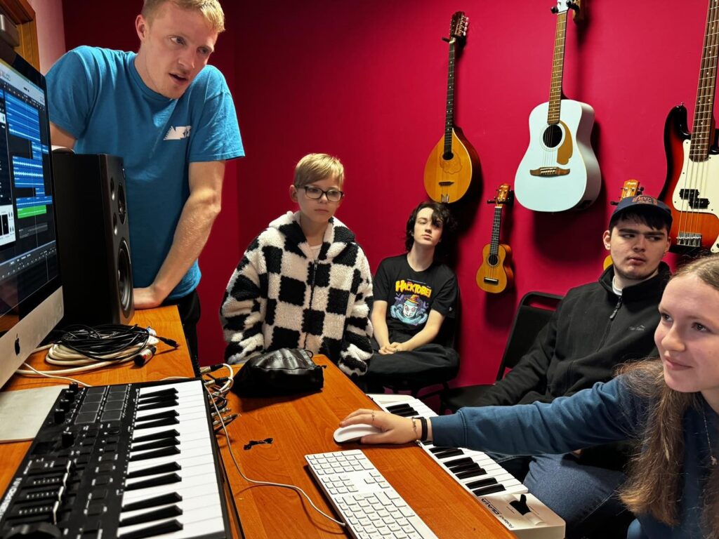 Five people are in a music studio with red walls, looking at a computer screen. Musical instruments, including guitars, hang on the wall. A keyboard and mouse are on the desk along with a MIDI controller.