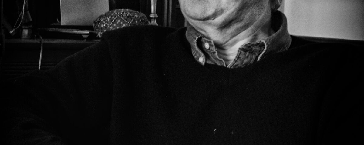 Black and white photo of an older man with gray hair sitting indoors, wearing a dark sweater over a collared shirt, looking thoughtfully to the side. Two table lamps and framed pictures are visible in the background.
