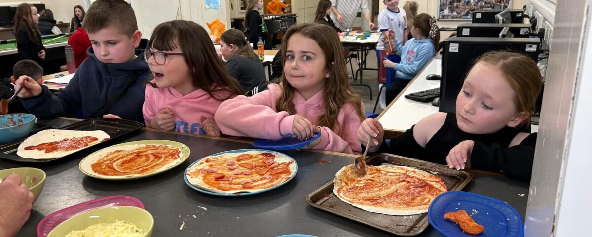 Four children sit at a counter spreading tomato sauce on pizza bases. Colorful bowls of toppings like cheese, onions, and peppers are in front of them. Other children and adults are seen in the background of a classroom.